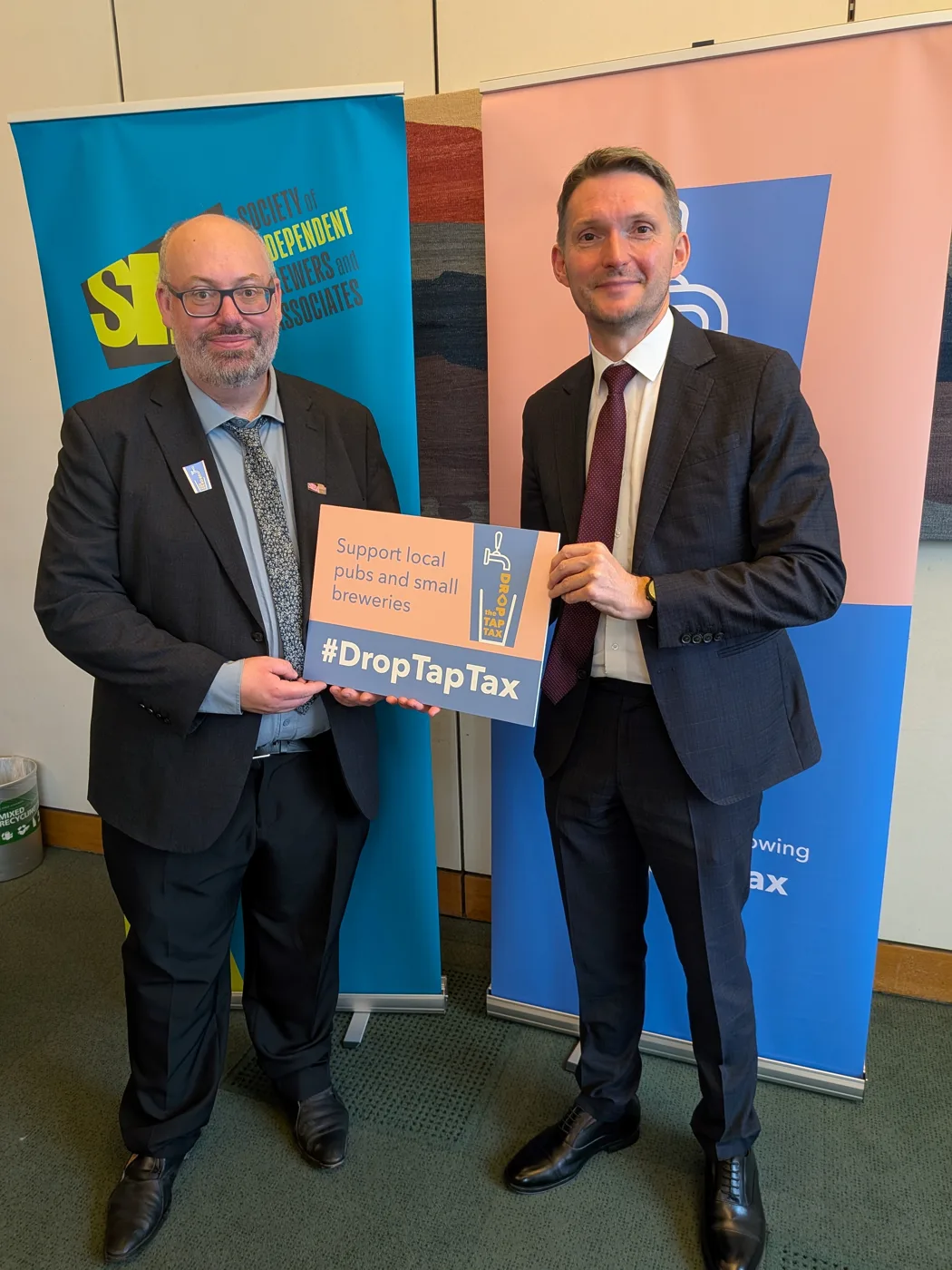 Chris Ward MP and a man in a dark suit holding a sign reading Support local pubs and small breweries, #DropTapTax, in front of SIBA banners