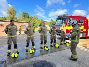 A row of recruits in fire kit with helmets at their feet, listening to an instructor holding a clipboard, with a fire engine behind