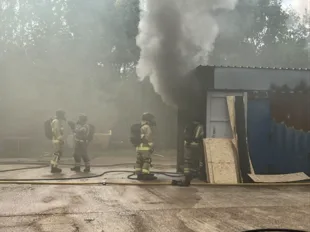 Firefighters in breathing apparatus beside a training unit with thick grey smoke pouring from its doorway