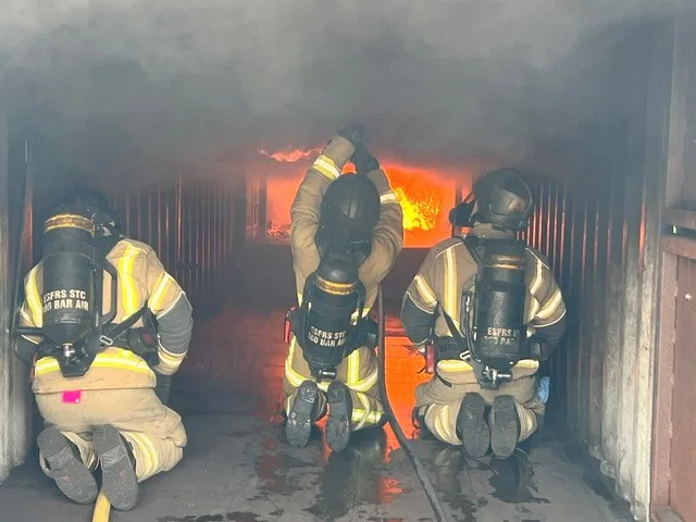 Three firefighters in breathing apparatus kneeling in a smoke-filled training unit, facing flames