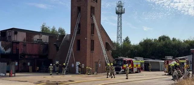 Firefighters pitching ladders against a brick training tower, with two fire engines parked nearby
