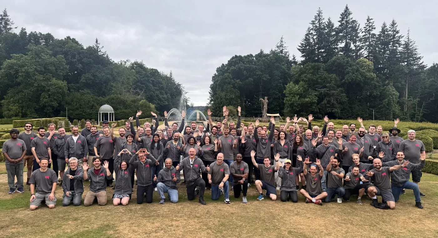 A large group in matching grey tops raise their hands on a lawn in front of a fountain.